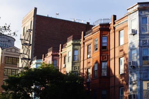A row of brick rowhouses with fire escapes and bay windows lit by warm low evening sun.