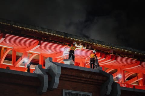 Workers on scaffolding tending the brightly lit red eaves of a temple roof at night.
