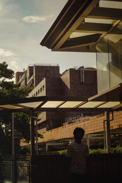 A person standing under a glass canopy with concrete campus buildings and trees behind.