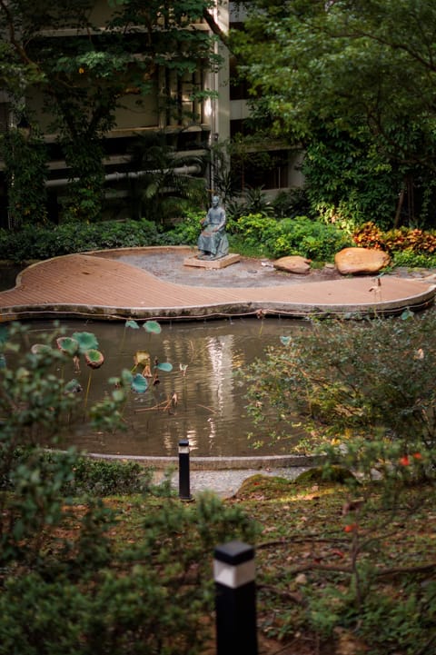 A small garden pond with lily pads and a seated statue, an apartment building rising behind.