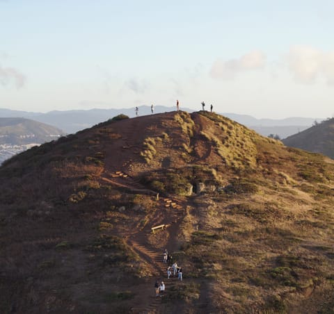 Hikers gathered on a grassy ridgeline hilltop at sunset, with distant hills beyond.