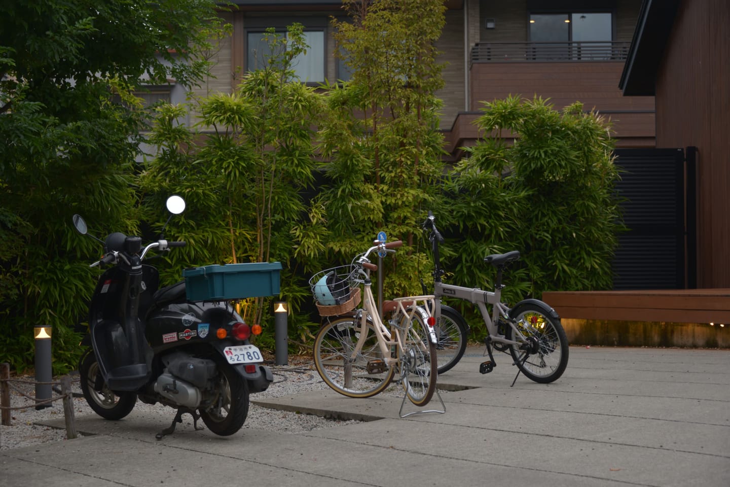 A moped and two bicycles parked beside bamboo outside a building at dusk.