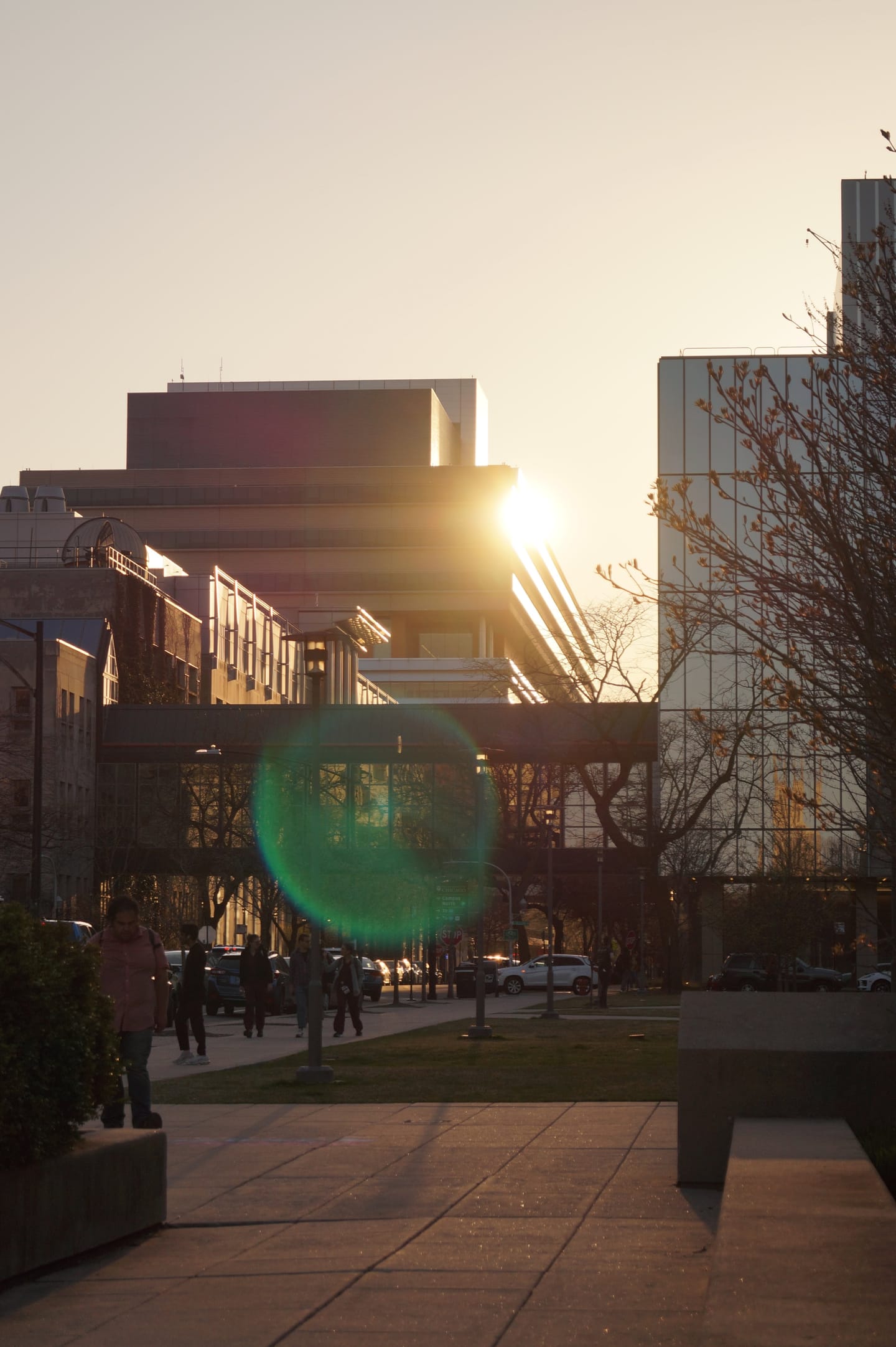 The sun flaring between modern campus buildings at sunset, with people crossing a plaza.