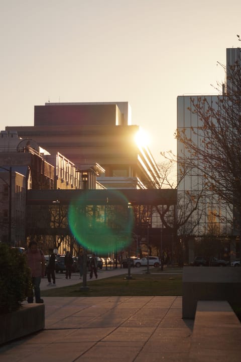 The sun flaring between modern campus buildings at sunset, with people crossing a plaza.