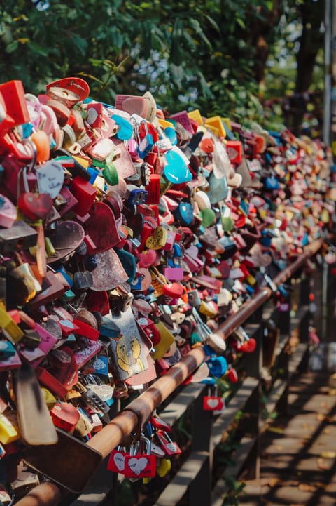 A fence densely covered with colourful love padlocks, sunlit trees behind.
