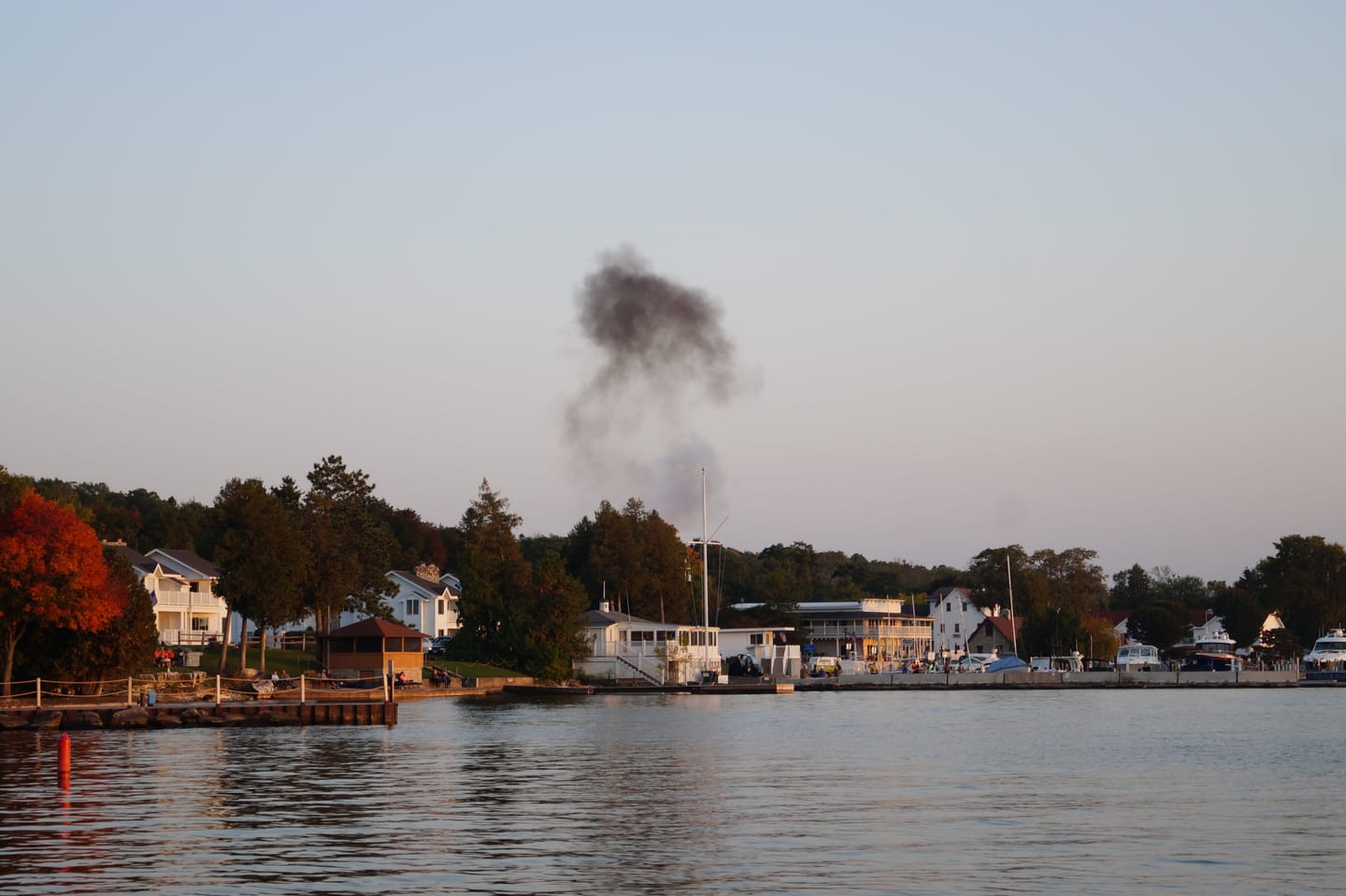 A lakeside town at dusk with a plume of dark smoke rising, autumn trees and calm water.