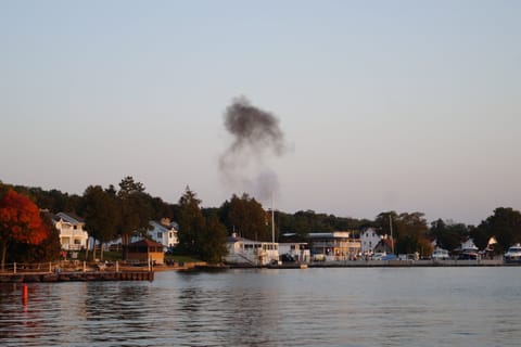A lakeside town at dusk with a plume of dark smoke rising, autumn trees and calm water.