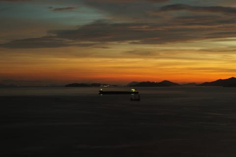 A cargo ship anchored at sea under a deep orange dusk sky, distant mountains on the horizon.