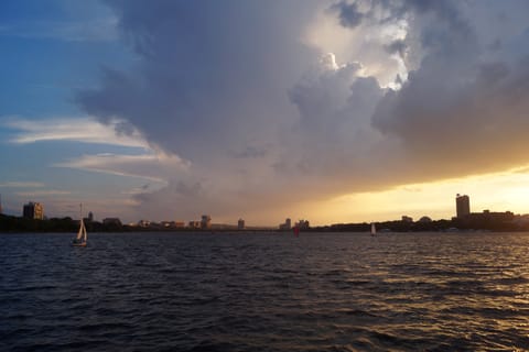 Sailboats on a lake at sunset beneath dramatic storm clouds, a skyline on the shore.
